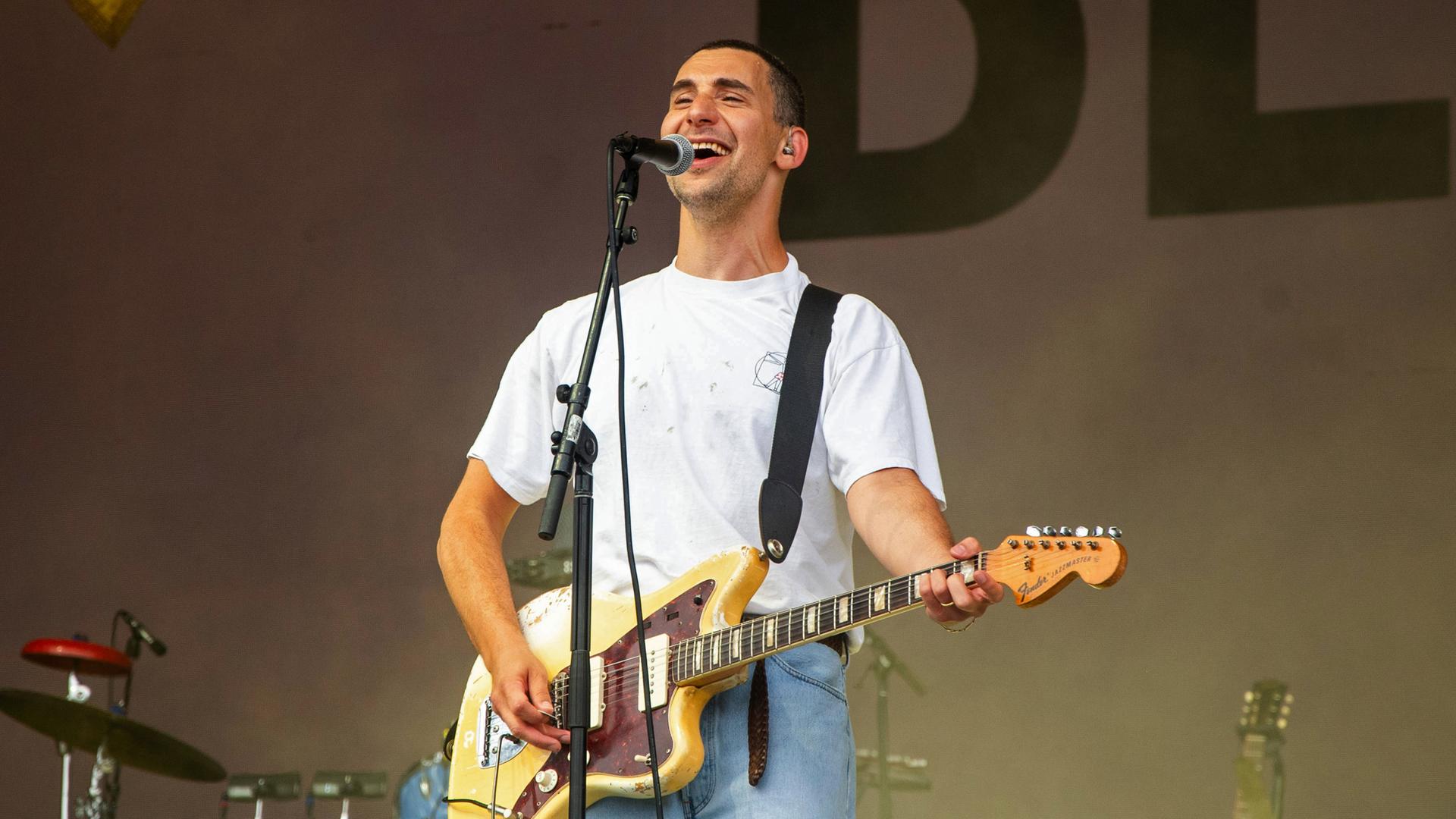 Ein junger Mann steht auf einer Bühne vor einem Mikrofon. Er singt und lacht ins Mikro und spielt dabei auf einer Gitarre, die er umhängen hat.