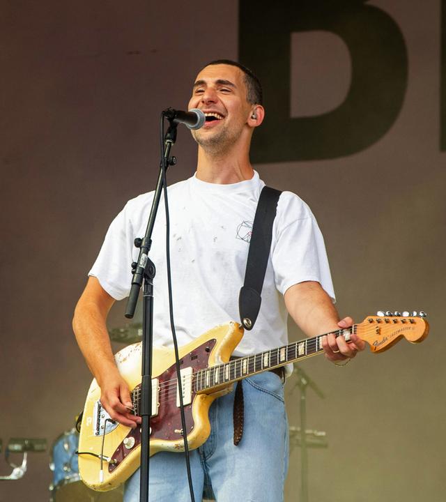 Ein junger Mann steht auf einer Bühne vor einem Mikrofon. Er singt und lacht ins Mikro und spielt dabei auf einer Gitarre, die er umhängen hat.