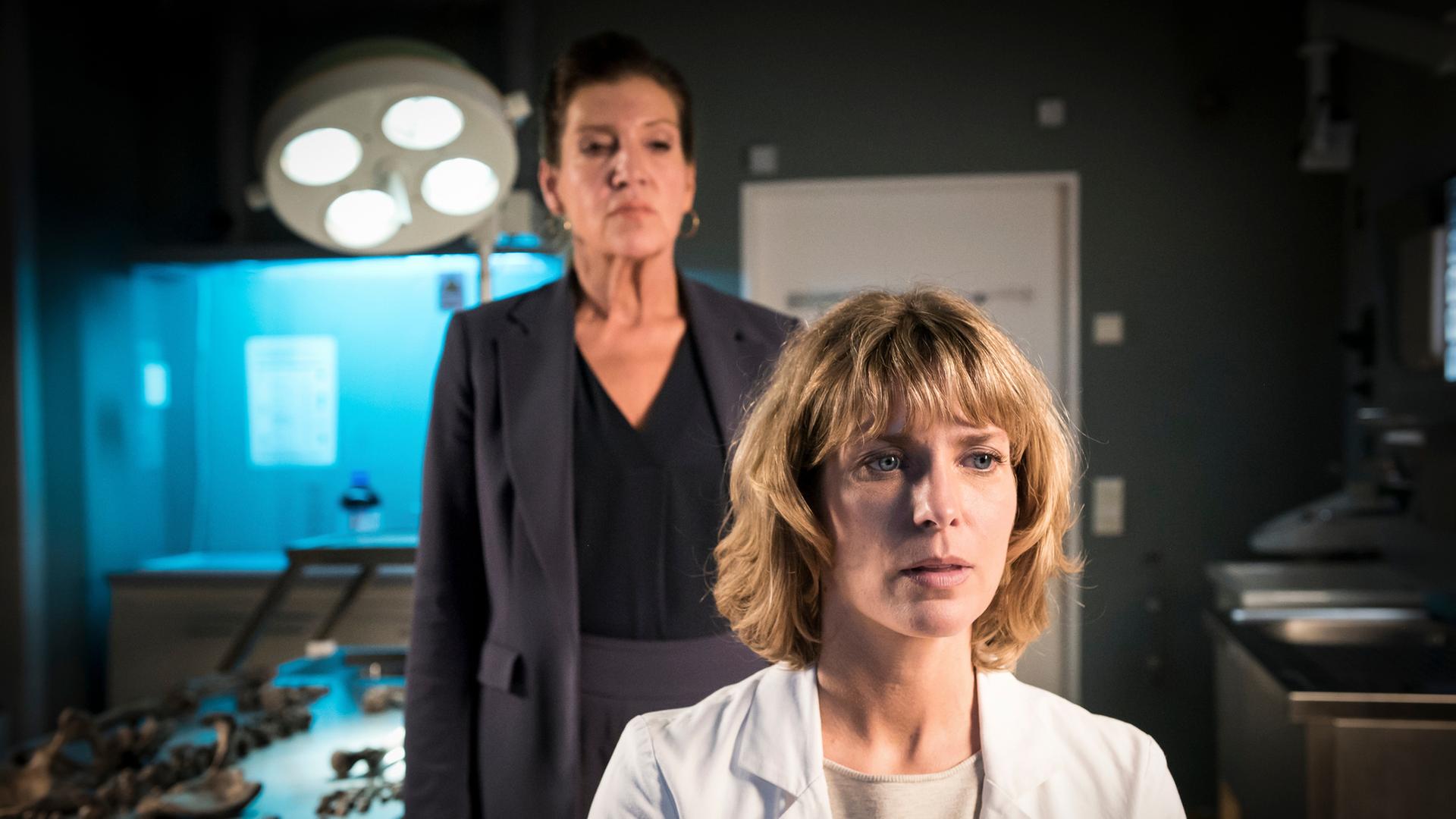 "Die Spezialisten - Sonne statt Reagan": Dr. Katrin Stoll (Valerie Niehaus) sitzt im Labor der IEK auf ihrem Stuhl und blickt vor sich. Hinter ihr steht Dr. Dorothea Lehberger (Katy Karrenbauer), die sie ernst ansieht.