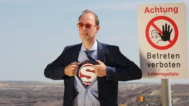 Martin Sonneborn steht vor einem "Achtung: Betreten verboten"-Schild und öffnet einen Teil seines Hemds, unter dem man ein großes S erkennt