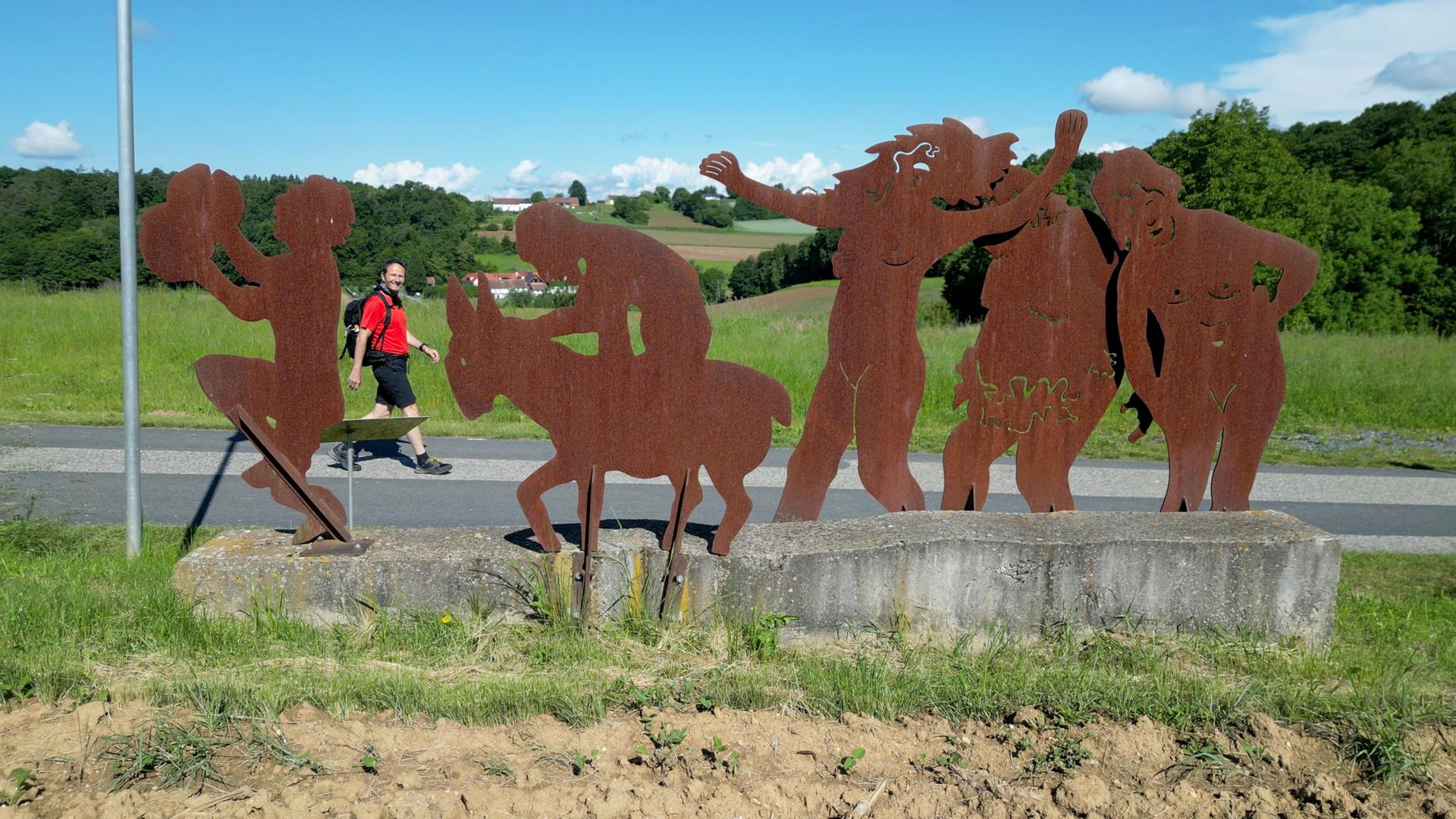 Das Bild zeigt eine Szene im Freien, in der ein Wanderer auf einem Weg entlanggeht. Der Wanderer trägt ein rotes Shirt, eine schwarze Hose und hat einen Rucksack auf dem Rücken. Neben dem Weg steht eine große, rustikale Skulpturengruppe aus Metall, die verschiedene Figuren darstellt. Diese Figuren sind in rostfarbener Farbe gehalten und scheinen verschiedene Aktivitäten zu zeigen, darunter eine Figur, die auf einem Esel sitzt und andere, die fröhlich wirken. Im Hintergrund sind sanfte grüne Hügel und ein blauer Himmel mit einigen Wolken zu sehen. Die Landschaft ist reichhaltig und bietet eine natürliche Kulisse für die dargestellte Szene.