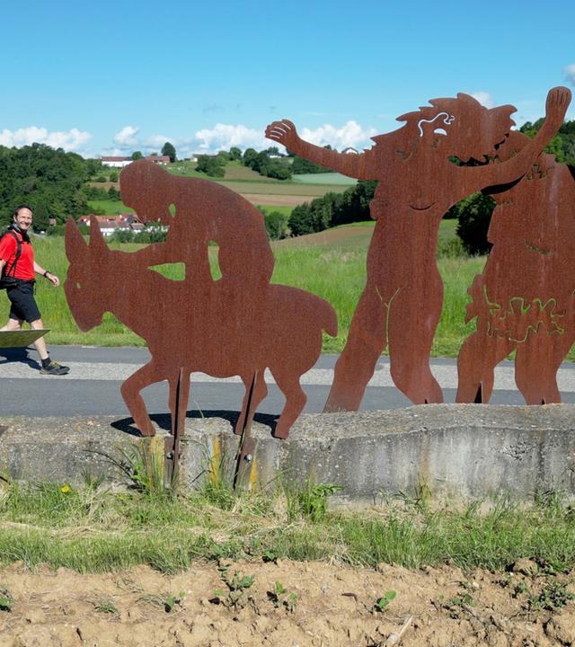 Das Bild zeigt eine Szene im Freien, in der ein Wanderer auf einem Weg entlanggeht. Der Wanderer trägt ein rotes Shirt, eine schwarze Hose und hat einen Rucksack auf dem Rücken. Neben dem Weg steht eine große, rustikale Skulpturengruppe aus Metall, die verschiedene Figuren darstellt. Diese Figuren sind in rostfarbener Farbe gehalten und scheinen verschiedene Aktivitäten zu zeigen, darunter eine Figur, die auf einem Esel sitzt und andere, die fröhlich wirken. Im Hintergrund sind sanfte grüne Hügel und ein blauer Himmel mit einigen Wolken zu sehen. Die Landschaft ist reichhaltig und bietet eine natürliche Kulisse für die dargestellte Szene.