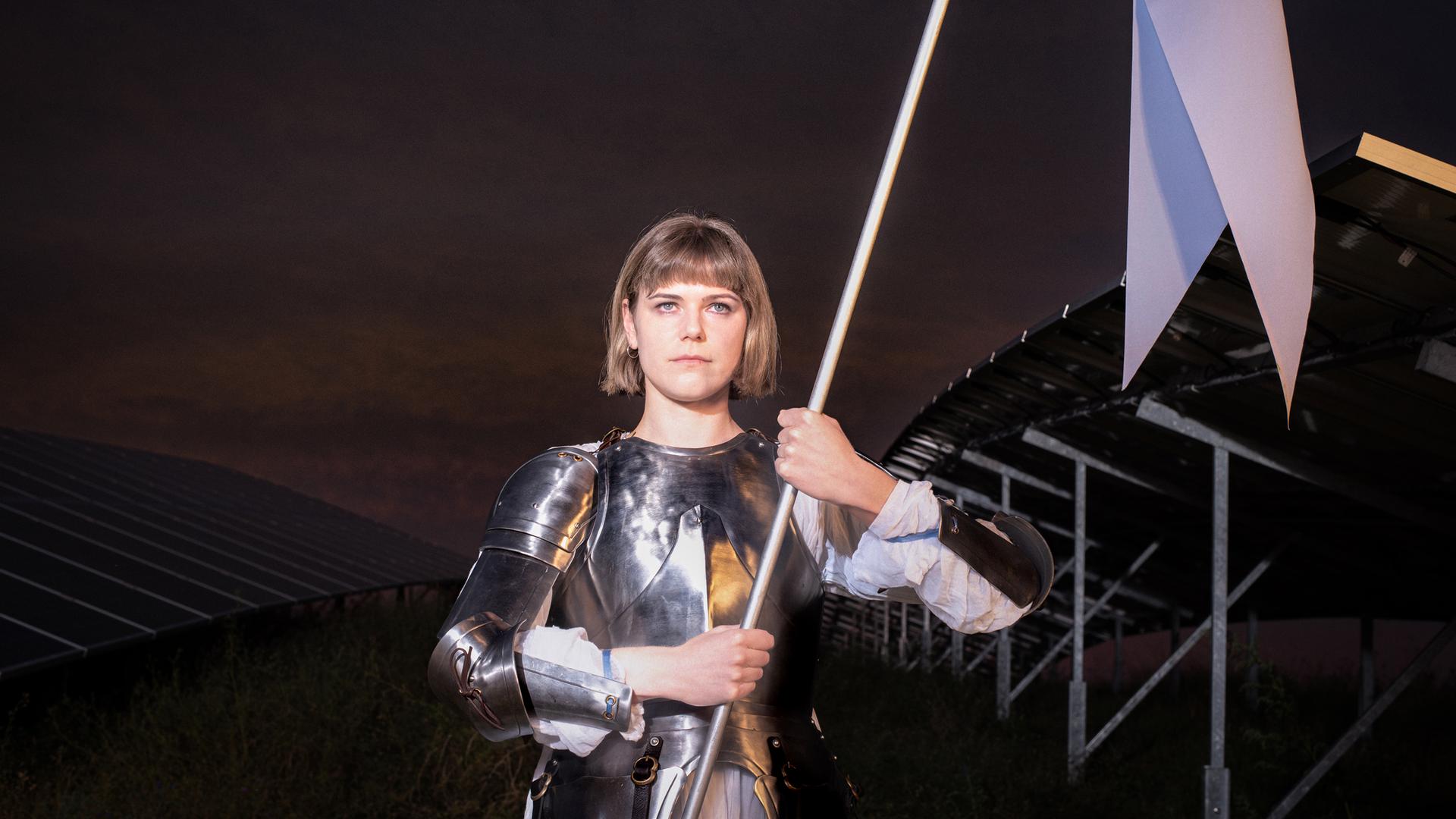 Johanna (Llewellyn Reichman) steht in Rüstung auf einem Feld mit Solarpaneelen. Sie trägt einen weißen Gambeson unter silbernen Metallplatten und hält eine weiße Fahne mit beiden Händen. Hinter ihr ist die Konstruktion der Solaranlage erkennbar, der Himmel wirkt dunkel und dramatisch. 