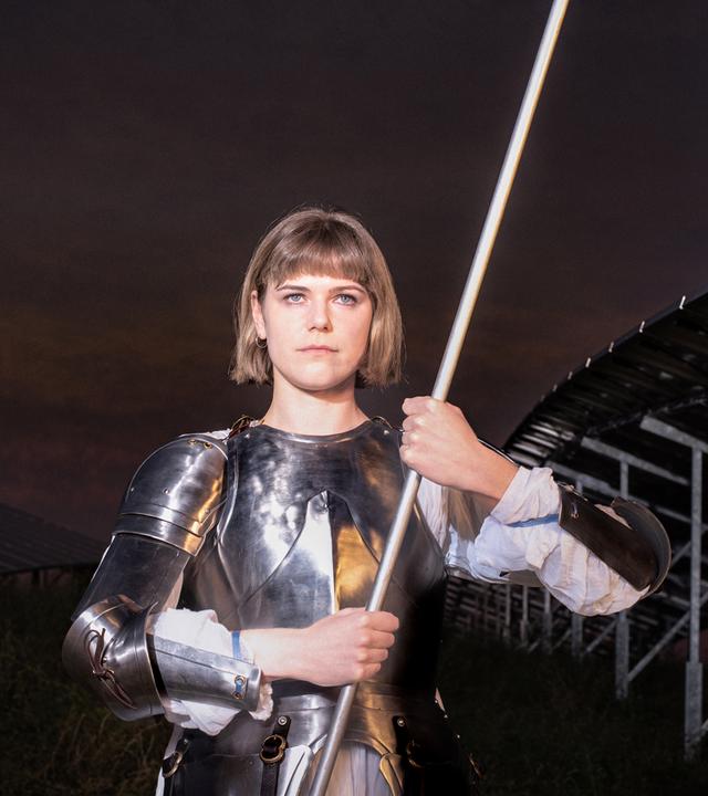 Johanna (Llewellyn Reichman) steht in Rüstung auf einem Feld mit Solarpaneelen. Sie trägt einen weißen Gambeson unter silbernen Metallplatten und hält eine weiße Fahne mit beiden Händen. Hinter ihr ist die Konstruktion der Solaranlage erkennbar, der Himmel wirkt dunkel und dramatisch. 