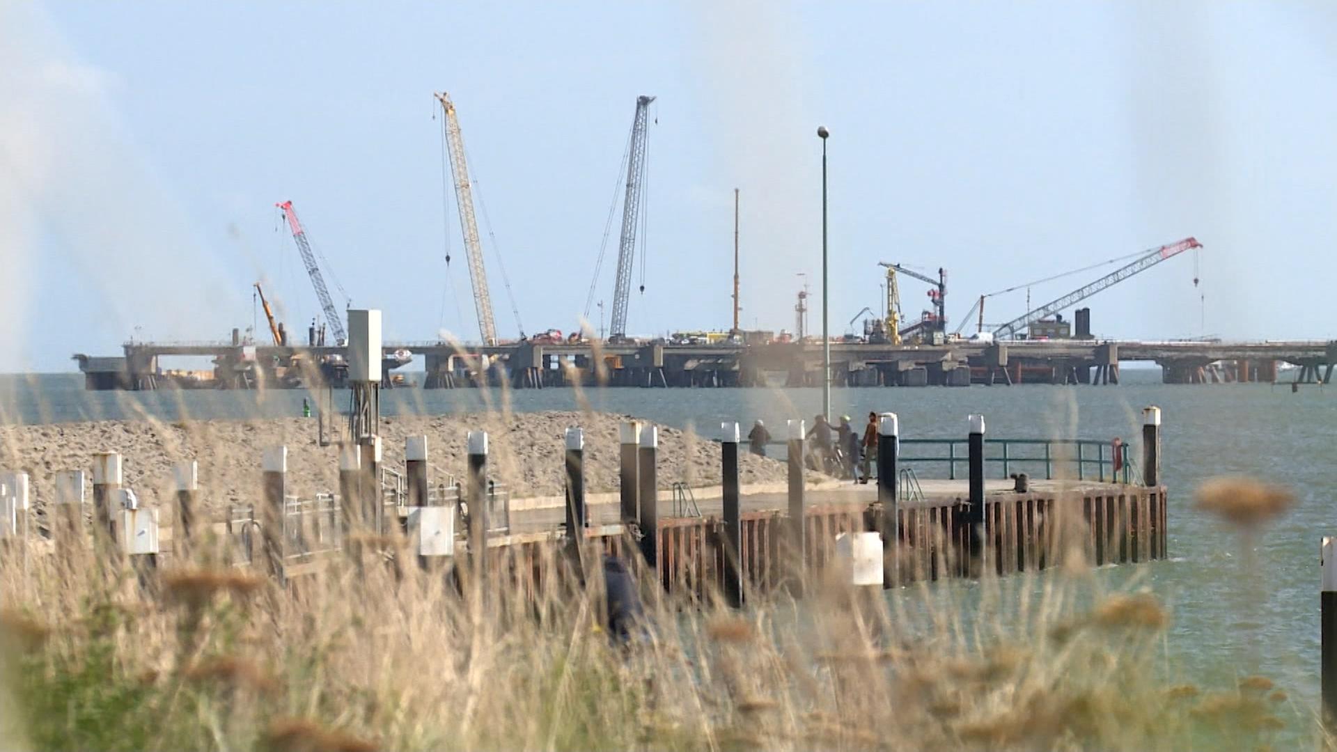 Im Vordergrund sind Gräser, das Meer und ein Steg zu sehen, im Hintergrund die Baustelle des LNG-Terminals im Wasser. 