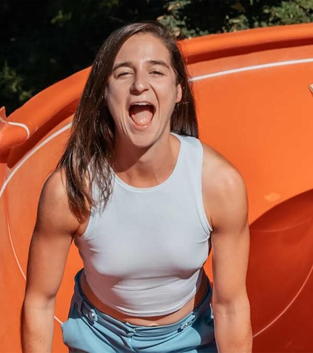 Das Bild zeigt eine junge Frau, die auf einer Wasserrutsche steht. Sie trägt ein helles, ärmelloses Oberteil und eine hellblaue Shorts. Die Frau hat einen lächelnden Ausdruck und scheint zu schreien oder zu lachen. Der Hintergrund ist grün und zeigt eine bewaldete Umgebung. Die Wasserrutsche, auf der sie steht, ist orange und hat eine spiralförmige Form. Es gibt keinen sichtbaren Wasserspieleffekt, und der Fokus liegt auf der positiven Ausstrahlung der Frau.