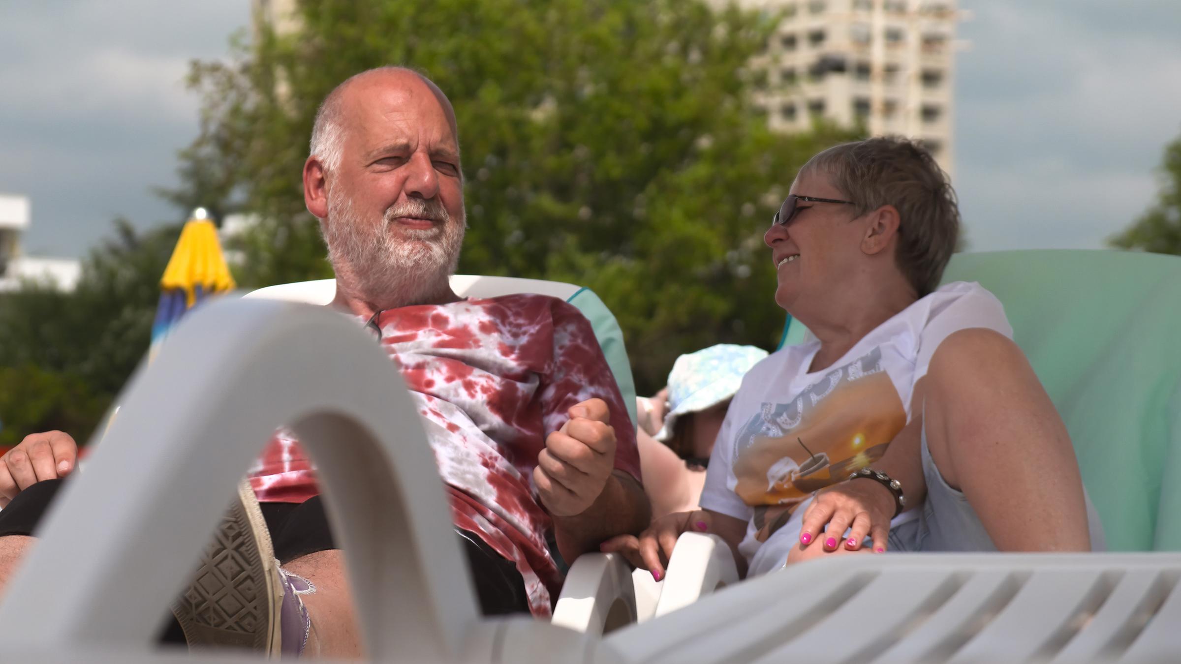 Kurt (57) und Verena (61) sitzen am Strand auf weißen Liegestühlen. Hinter ihnen stehen blau-gelbe Sonnenschirme vor Hotelfassaden.
