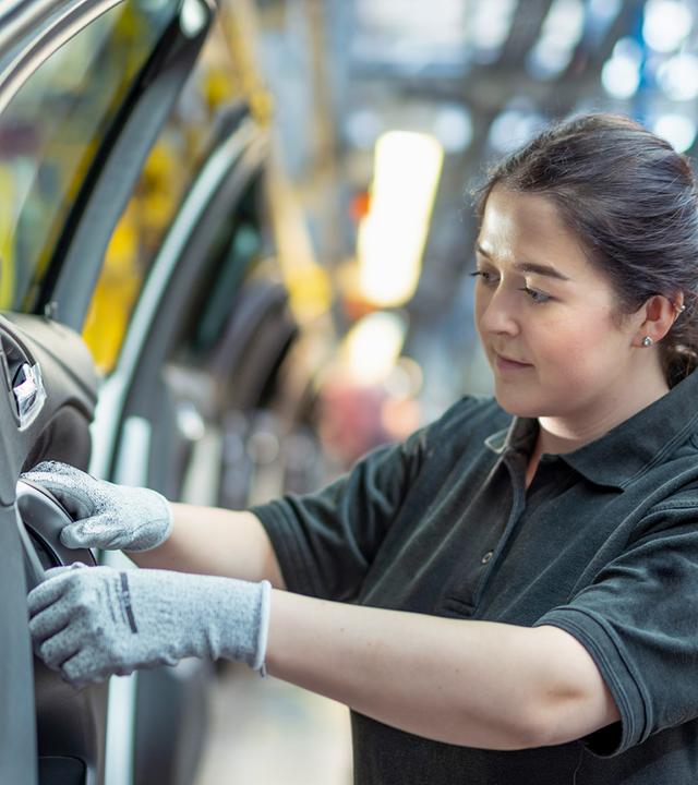 Im Vordergrund ist die geöffnete Tür eines hellen Fahrzeugs zu sehen. Rechts daneben arbeitet eine Beschäftigte mit behandschuhten Händen am Bereich des inneren Türgriffs. Sie blickt auf die Stelle, an der sie arbeitet. Dahinter sind Teile einer Produktionshalle erkennbar.