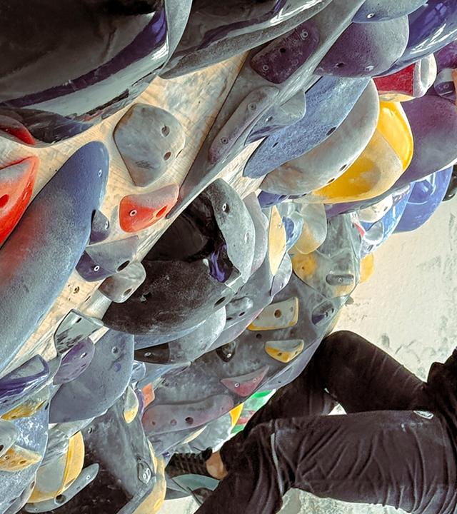 Eine Person klettert an einer mit bunten, unterschiedlichen Griffen versehenen Kletterwand. Die Kletterin trägt ein ärmelloses Oberteil und schwarze Hose.