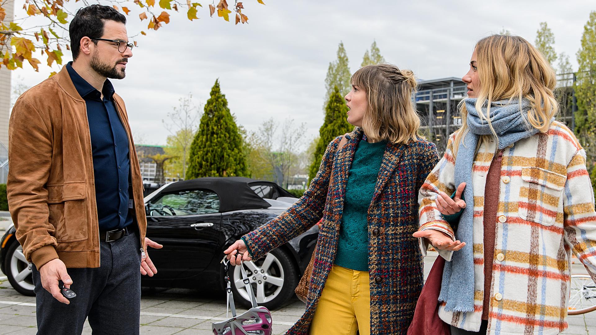 Dr. Tom Koopmann (Tobias Licht) steht auf dem Krankenhausparkplatz Rike (Isabell Horn) und Betty (Annina Hellenthal) provokativ schauend gegenüber. Beide Frauen schauen fragend zurück.