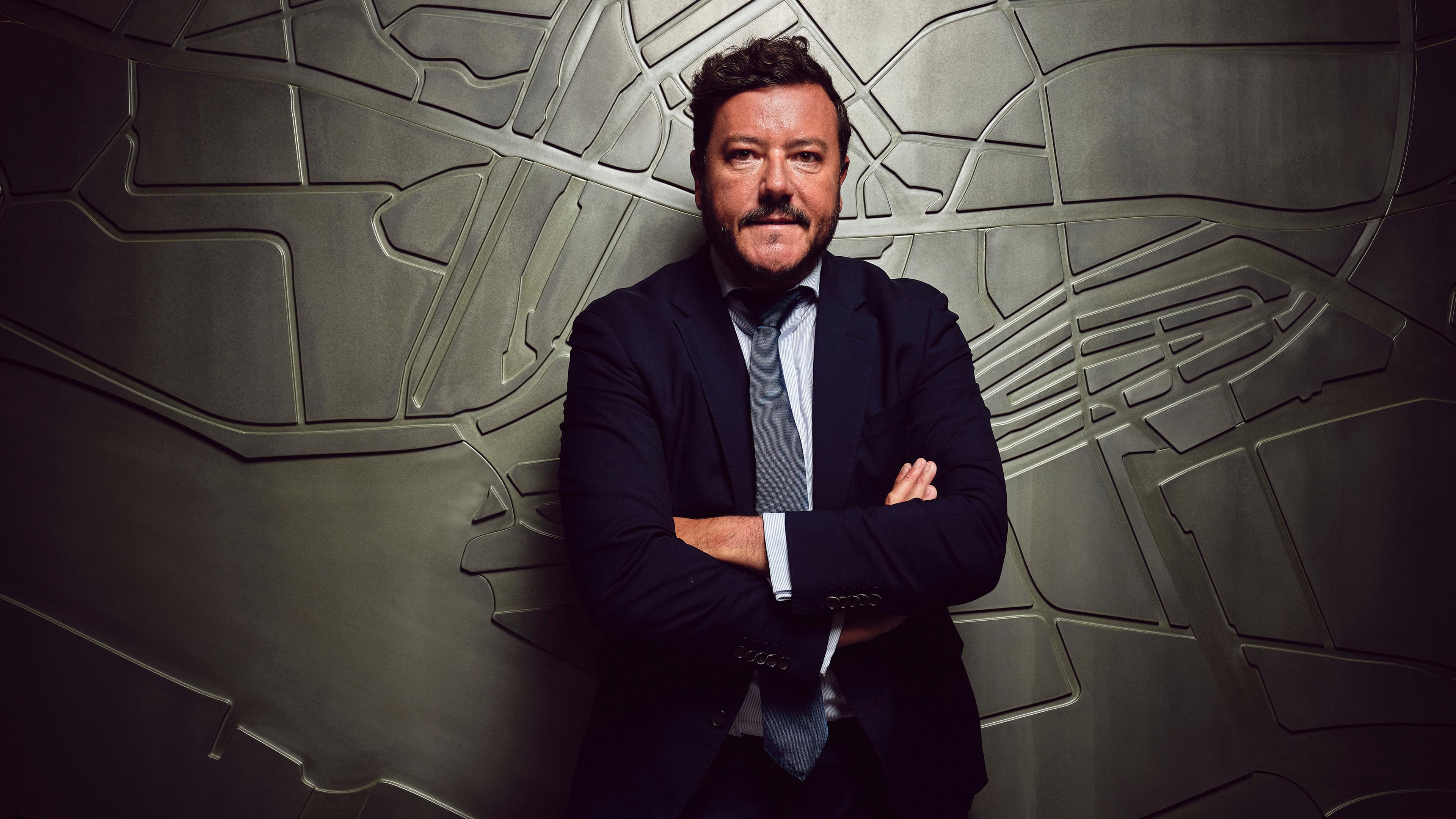 Rene Benko steht mit verschränkten Armen im dunklen Anzug vor einer Wand mit einem silberfarbenen Relief. Er schaut ernst in die Kamera.