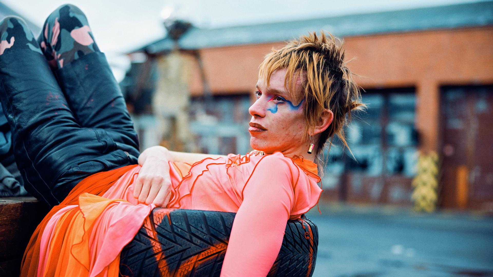 Das Bild zeigt eine Person, die lässig auf einem Stapel Reifen liegt. Die Person trägt ein auffälliges, farbenfrohes Outfit mit einem leuchtend pinken Oberteil und einer orange-pinken, durchscheinenden Rock-ähnlichen Schicht. Die Hose ist dunkel mit einem Tarnmuster. Das Gesicht der Person ist markant geschminkt, mit blauen Akzenten um das rechte Auge und roten Lippen. Die Haare sind kurz und wild gestylt, in einem rötlich-blonden Ton. Im Hintergrund ist eine unscharfe städtische Umgebung mit Gebäuden und einem leicht bewölkten Himmel zu sehen.