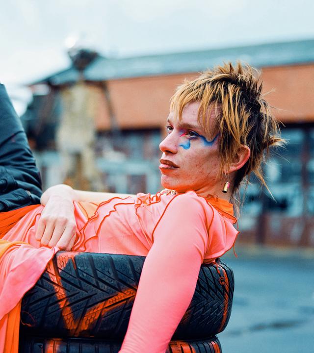 Das Bild zeigt eine Person, die lässig auf einem Stapel Reifen liegt. Die Person trägt ein auffälliges, farbenfrohes Outfit mit einem leuchtend pinken Oberteil und einer orange-pinken, durchscheinenden Rock-ähnlichen Schicht. Die Hose ist dunkel mit einem Tarnmuster. Das Gesicht der Person ist markant geschminkt, mit blauen Akzenten um das rechte Auge und roten Lippen. Die Haare sind kurz und wild gestylt, in einem rötlich-blonden Ton. Im Hintergrund ist eine unscharfe städtische Umgebung mit Gebäuden und einem leicht bewölkten Himmel zu sehen.