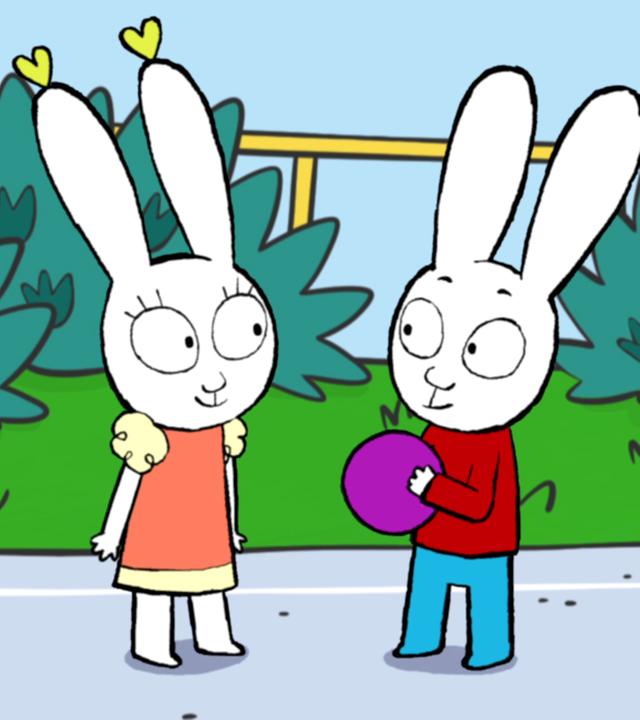 Simon hält einen lila Ball in seiner Hand und steht Lou gegenüber, die ihn mit großen Augen ansieht. 