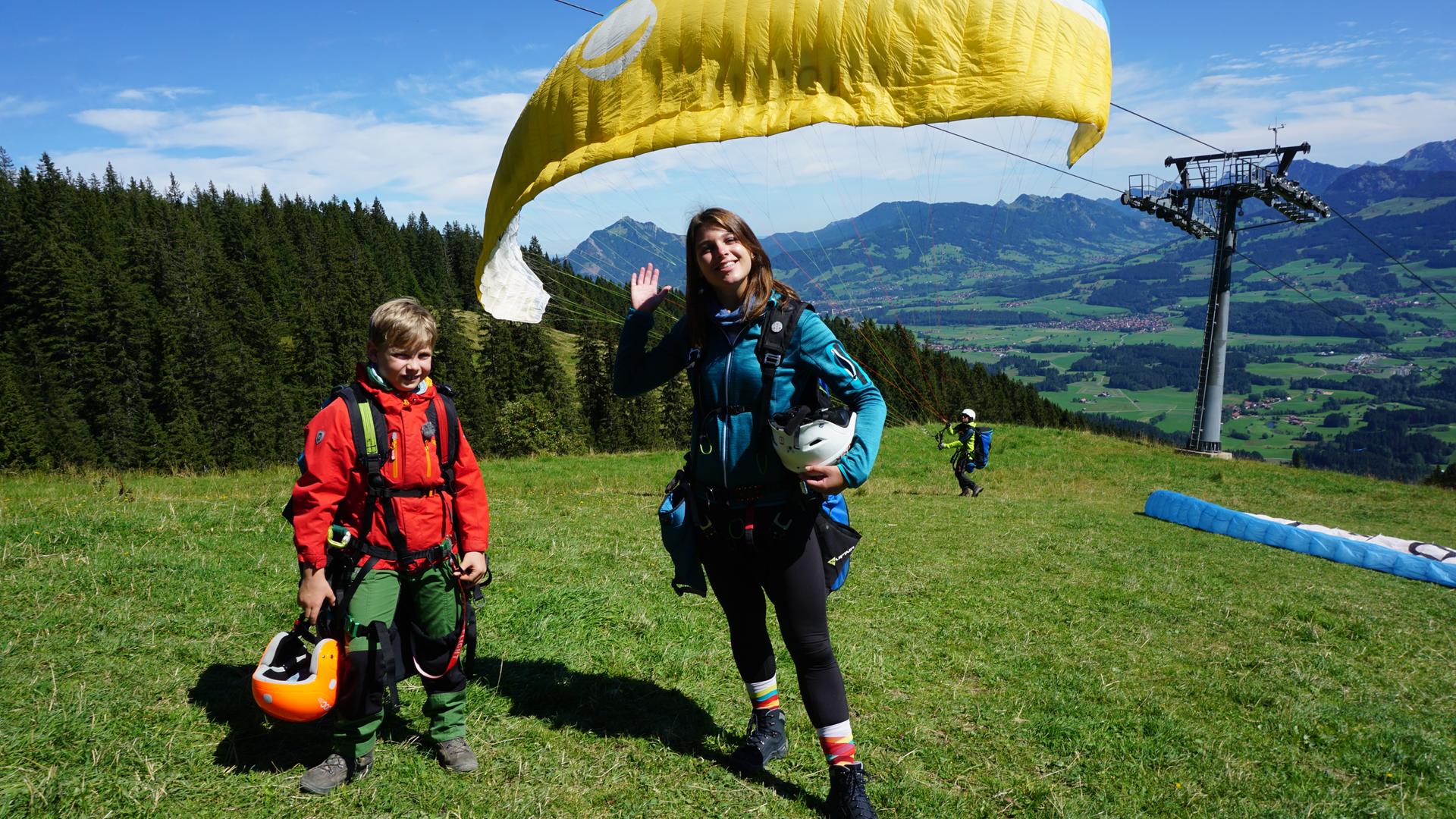 Diego und die winkende Laura stehen oben auf einem Berg. Hinter ihnen schwebt der Gleitschirm.