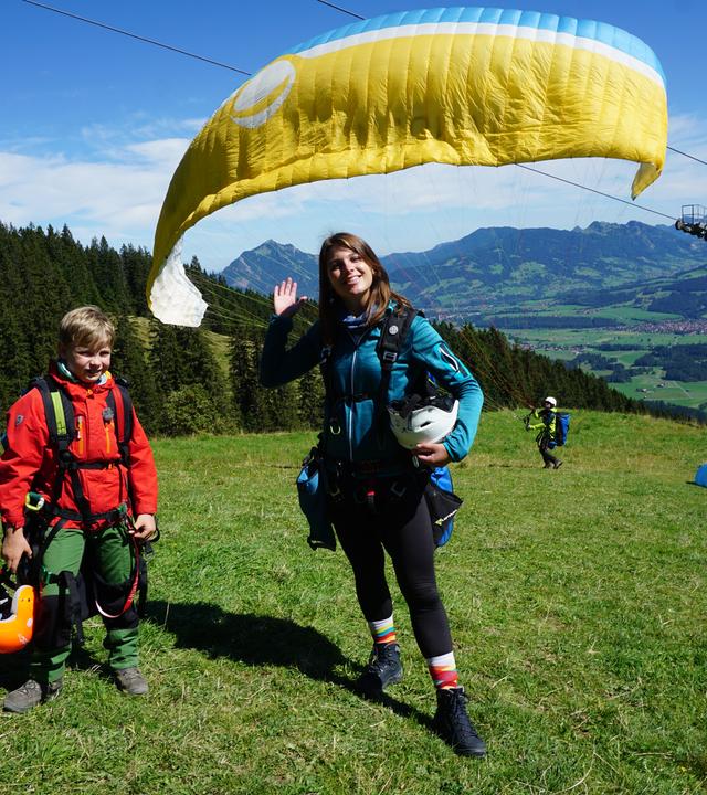 Diego und die winkende Laura stehen oben auf einem Berg. Hinter ihnen schwebt der Gleitschirm.
