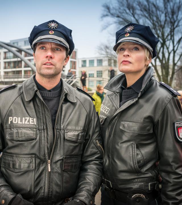 "Notruf Hafenkante - Sprache der Stärke": Die Polizeibeamten Mattes Seeler (Matthias Schloo) und Melanie Hansen (Sanna Englund) stehen nebeneinander und schauen auf etwas vor sich. Im Hintergrund steht ihr Streifenwagen hinter einem Absperrband vor dem Menschen stehen.