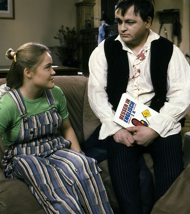 "Sprechstunde bei Dr. Frankenstein - English for Monsters": Franzi (Floriane Eichhorn) und Lenni (Michael Trischan) sitzen in einem Wohnraum auf dem Sofa und schauen sich an. Lenni hält ein Buch mit dem Titel "Besser in Englisch" in der Hand.