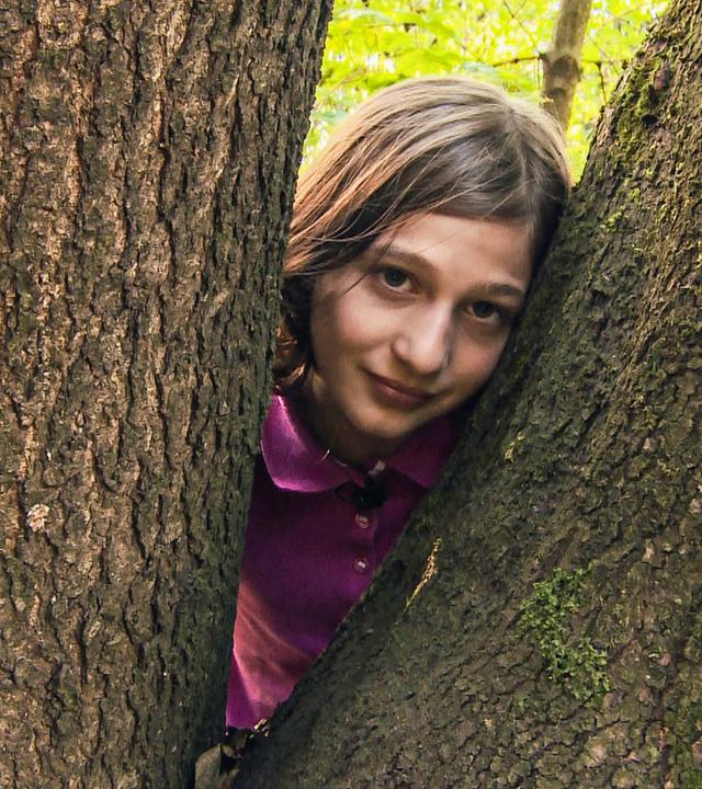 Yael schaut zwischen zwei riesigen Baumstämmen in die Kamera.