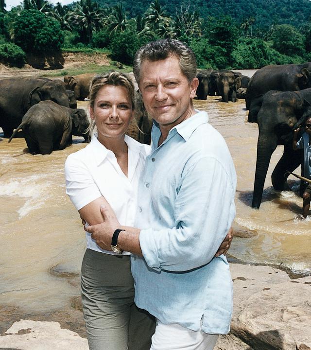 "Das Traumschiff - Sri Lanka": Ina (Janette Rauch) und Klaus (Miroslav Nemec) stehen Arm in Arm am Fluss und lächeln in die Kamera. Im Hintergrund steht eine Elefantenherde im Fluss, ein Elefant wird von einem Elefantenführer geleitet.
