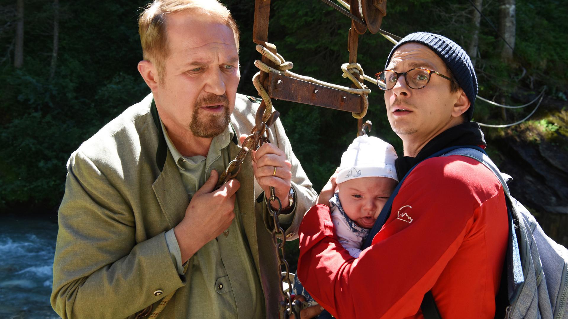 Ein Mann in roter Jacke mit dem Baby auf dem Arm. Und ein Mann der einen Gurt eines Liftes festhält.