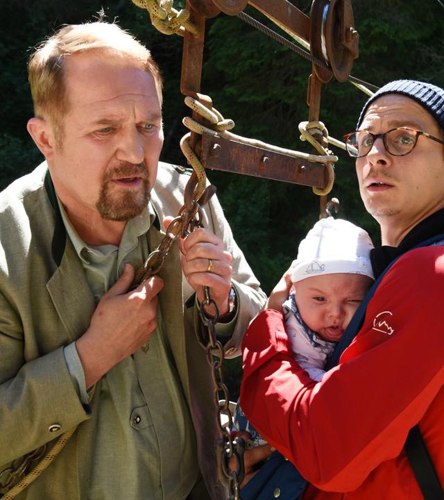 Ein Mann in roter Jacke mit dem Baby auf dem Arm. Und ein Mann der einen Gurt eines Liftes festhält.