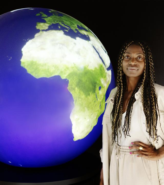 Christiana steht vor einem schwarzen Hintergrund, neben ihr ist ein Globus zu sehen.