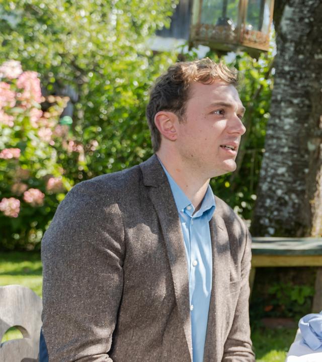 Kommissar Schmidt (Moritz von Zeddelmann) sitzt am Frühstückstisch im Garten und sieht Gerlinde Meyr (Ulla Geiger) an, die am Tisch steht und ihm lächelnd Kaffee einschenkt.