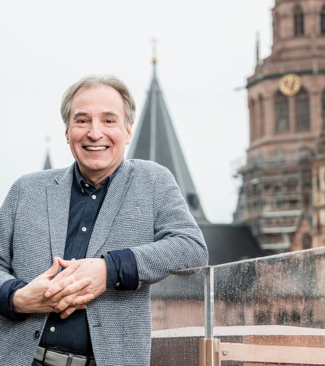 Lächelnder Mann in grauem Tweed-Sakko mit gefalteten Händen, im Hintergrund Turmspitzen einer Kirche.