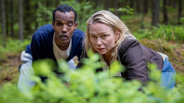 "Detective No. 24: Anna": Tilda (Malin Levanon) und Ibraahin (Nasir Farah Dhagole) knien im Wald hinter einem Gebüsch. Sie wollen nicht gesehen werden.