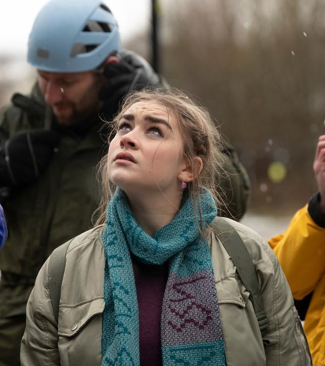 Morgan Strandhed (Silvie Furneaux) blickt mit Sorgenfalten auf der Stirn skeptisch nach oben. Um den Hals trägt sie Sigis Schal. Um sie herum stehen Kletterer (Komparsen), die ihre Helmgurte schließen.