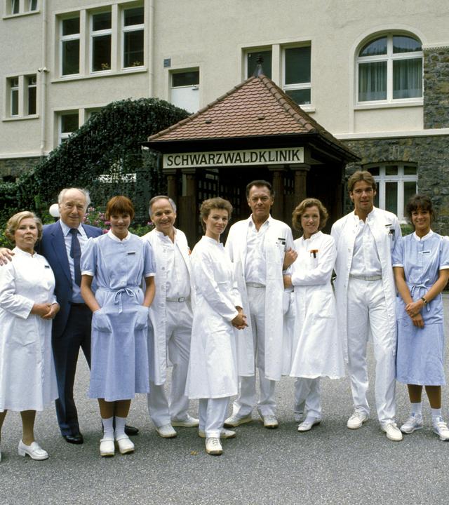 "Die Schwarzwaldklinik - Die Heimkehr (1)": Oberschwester Hildegard (Eva Maria Bauer), Verwaltungsdirektor Alfred Mühlmann (Alf Marholm), Schwester Ina (Gaby Fischer), Dr. Gerd Wolter (Franz Rudnick), Dr. Katarina Gessner (Ilona Grübel), Prof. Klaus Brinkmann (Klausjürgen Wussow), Schwester Christa (Gaby Dohm), Dr. Udo Brinkmann (Sascha Hehn) und Schwester Elke (Barbara Wussow) stehen nebeneinander vor der Schwarzwaldklinik und lächeln Richtung Kamera.
