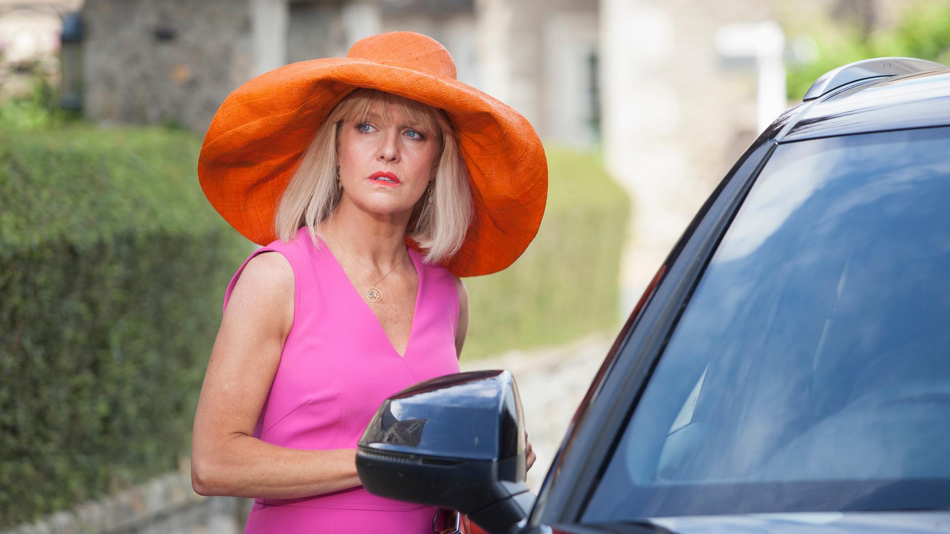 "Agatha Raisin - Der tote Friseur Teil 1": Agatha Raisin (Ashley Jensen) steht neben einem Auto und trägt ein auffälliges Outfit, das aus einem pinken Kleid und einem orangefarbenen Hut mit sehr breiter Krempe besteht.