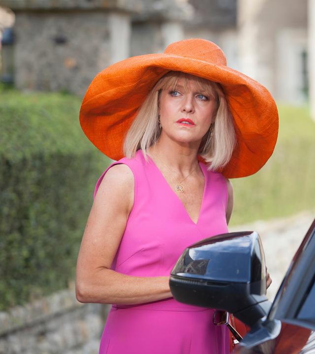 "Agatha Raisin - Der tote Friseur Teil 1": Agatha Raisin (Ashley Jensen) steht neben einem Auto und trägt ein auffälliges Outfit, das aus einem pinken Kleid und einem orangefarbenen Hut mit sehr breiter Krempe besteht.