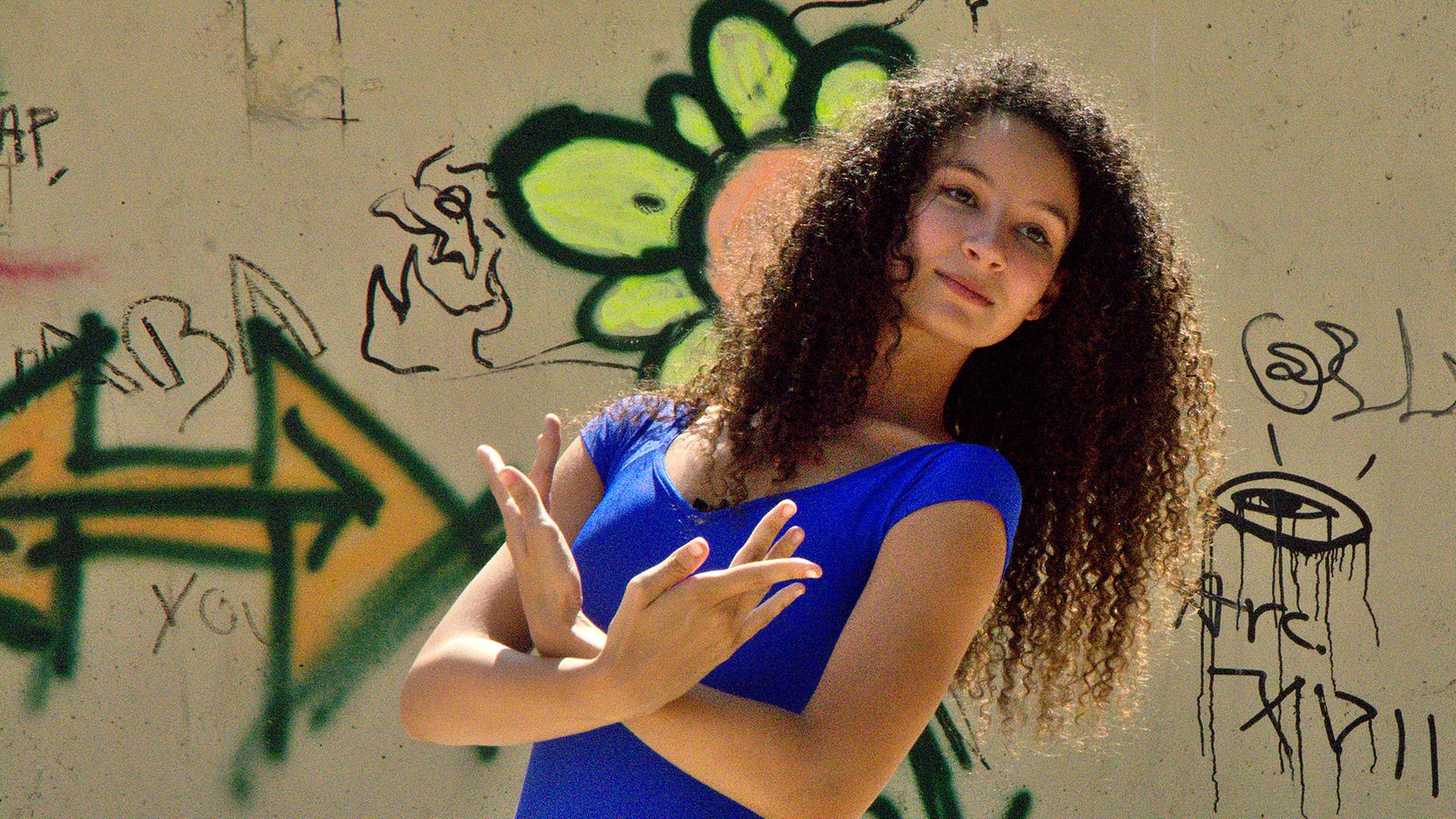 Ines (mittig) macht eine Ballettpose. Sie steht vor einer steinernen Wand mit Graffiti darauf.