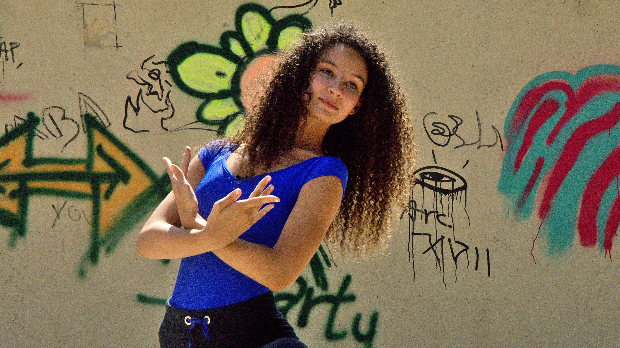 Ines (mittig) macht eine Ballettpose. Sie steht vor einer steinernen Wand mit Graffiti darauf.