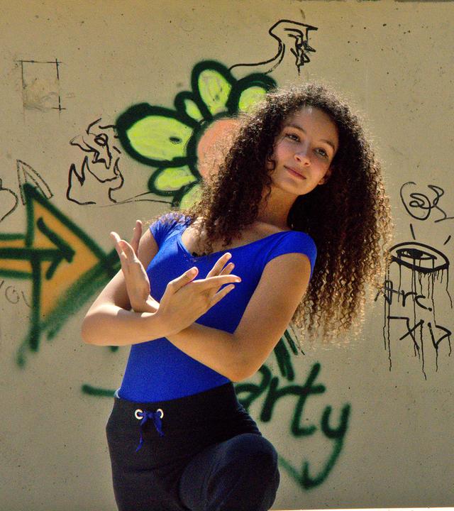 Ines macht eine Ballettpose. Sie steht vor einer steinernen Wand mit Graffiti darauf.