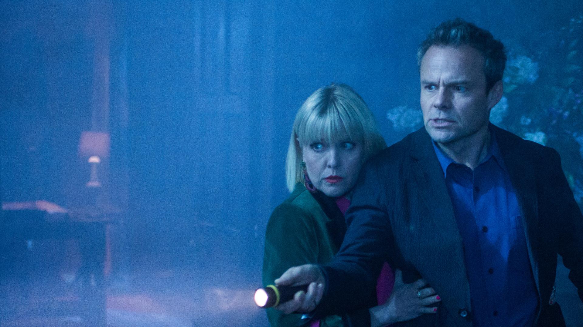 "Agatha Raisin – Und das Geisterhaus Teil 1": Agatha Raisin (Ashley Jensen) klammert sich ängstlich an James Lacey (Jamie Glover), der mit einer Taschenlampe ein dunkles Zimmer absucht.