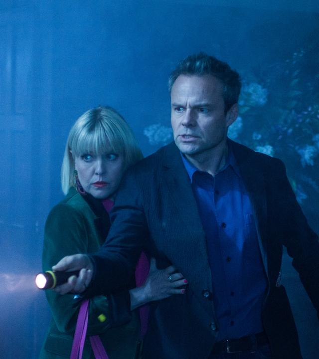 "Agatha Raisin – Und das Geisterhaus Teil 1": Agatha Raisin (Ashley Jensen) klammert sich ängstlich an James Lacey (Jamie Glover), der mit einer Taschenlampe ein dunkles Zimmer absucht.