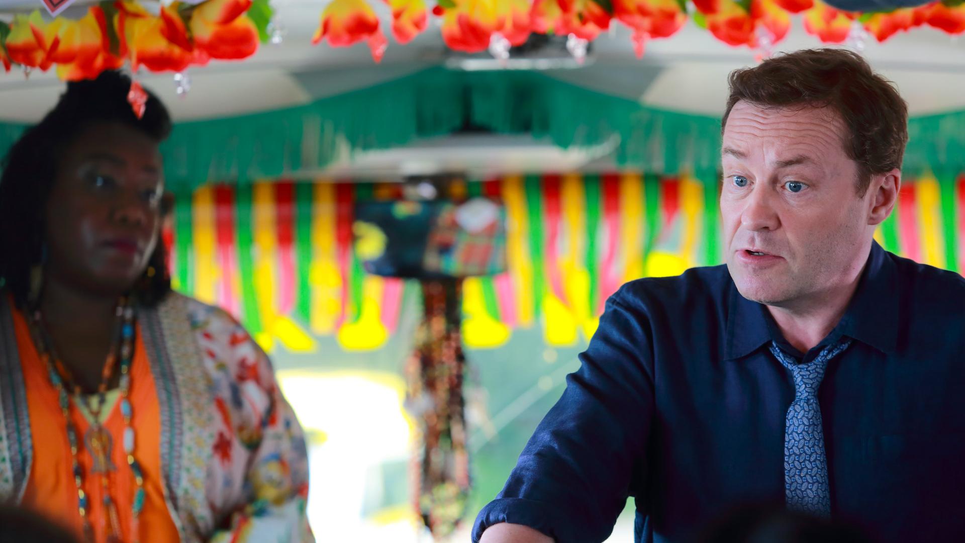 "Death in Paradise - Der Tote im Bus": DI Jack Mooney (Ardal O'Hanlon) und Butterfly Brown (Chizzy Akudolu) stehen in einem Bus nebeneinander und haben einen ernsten Blick.