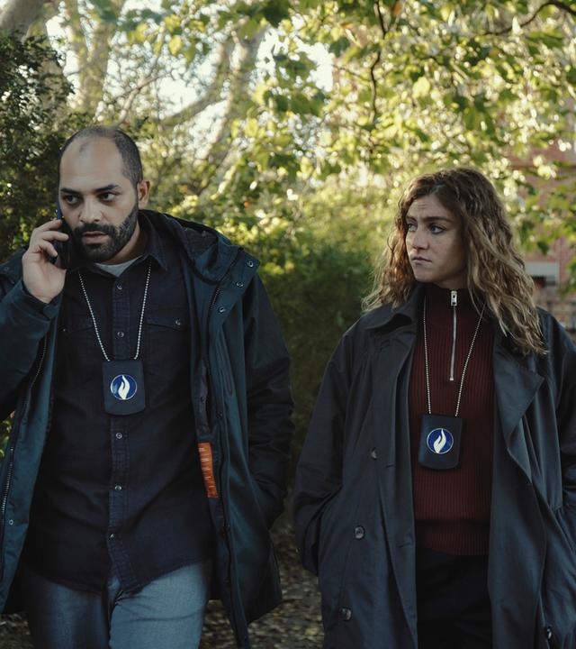 "Juliet - Die Tote im Kanal": Jamal (Nabil Mallat) und Juliet (Charlotte De Bruyne) gehen in einem Garten nebeneinander. Er telefoniert. Sie schaut in Jamals Richtung.