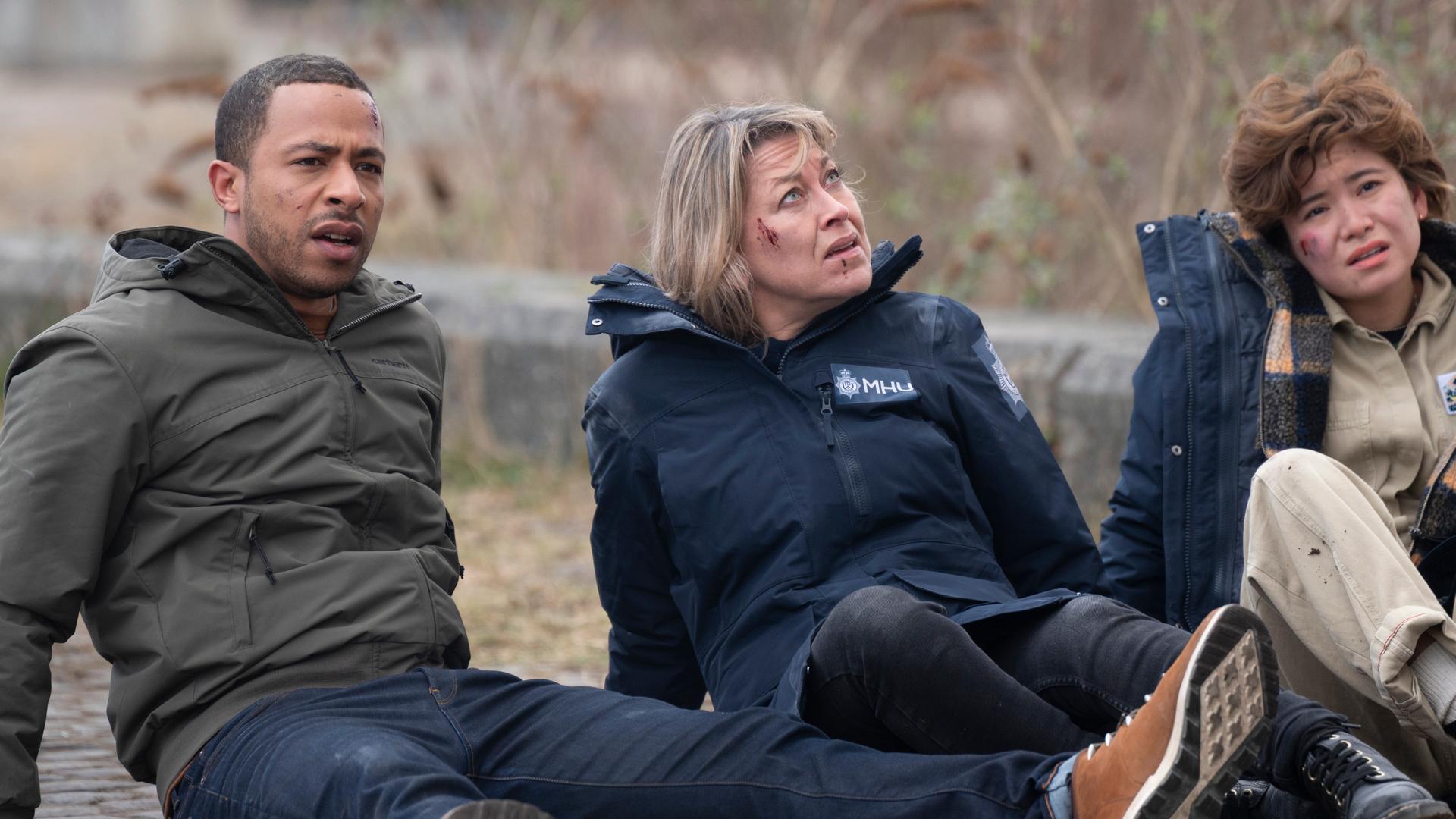 "Annika - Mord an Schottlands Küste - Familie": Tyrone Clarke (Ukweli Roach), Annika Strandhed (Nicola Walker) und Blair Ferguson (Katie Leung) sitzen im Freien auf dem Boden, sie haben Schrammen im Gesicht und blicken erschöpft.