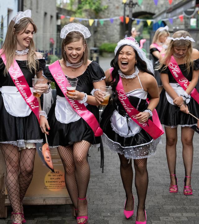 Sinead (Sorcha Fahy), Niamh (Juliette Crosbie) und Charlotte (Rhea Rose Rodillas) gehen nebeneinander durch eine Straße, in der mehrere Bars ansässig sind. Alle drei lachen. Holly (Ali Hardiman) bleibt etwas zurück und schaut nach unten. Noch weiter im Hintergrund steht Aisling (Sophie Vavasseur). Alle Frauen tragen Hausmädchen-Kostüme und pinkfarbene Schärpen.
