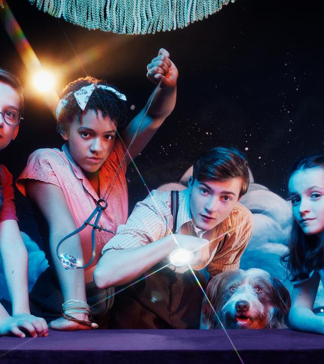 Dick (Kit Rakusen), George (Diaana Babnicova), Julian (Elliott Rose), Timmy (Kip) und Anne (Flora Jacoby Richardson) stehen vor einem Tisch auf einer Bühne und blicken in die Kamera. George schwingt ein Pendel mit einem Diamanten, während Julien mit einer Taschenlampe in die Kamera leuchtet.