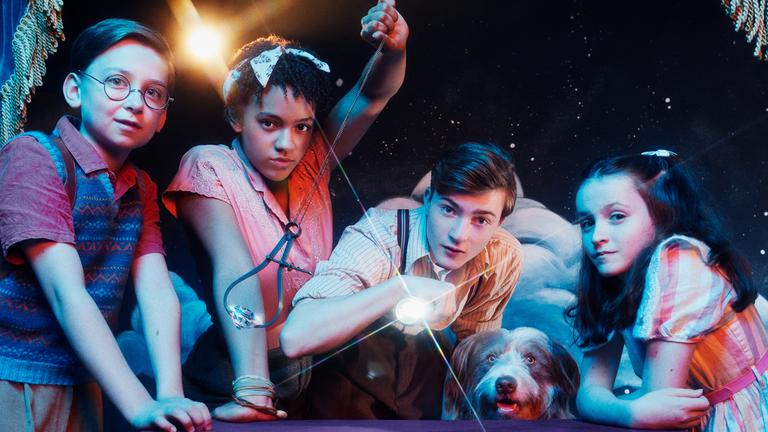 Dick (Kit Rakusen), George (Diaana Babnicova), Julian (Elliott Rose), Timmy (Kip) und Anne (Flora Jacoby Richardson) stehen vor einem Tisch auf einer Bühne und blicken in die Kamera. George schwingt ein Pendel mit einem Diamanten, während Julien mit einer Taschenlampe in die Kamera leuchtet.