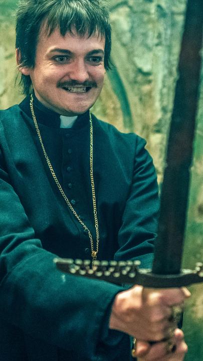 "Fünf Freunde - Der Fluch der Felseninsel": Thomas Wentworth (Jack Gleeson) steht in einer Kirche und hält ein Schwert in beiden Händen. Er ist wie ein Priester gekleidet. Neben ihm steht sein Assistent Franklin Boswell (William Abadie), der einen Anzug, eine Schiebermütze und Handschuhe trägt. Beide grinsen.
