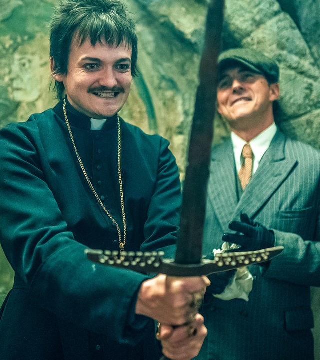 "Fünf Freunde - Der Fluch der Felseninsel": Thomas Wentworth (Jack Gleeson) steht in einer Kirche und hält ein Schwert in beiden Händen. Er ist wie ein Priester gekleidet. Neben ihm steht sein Assistent Franklin Boswell (William Abadie), der einen Anzug, eine Schiebermütze und Handschuhe trägt. Beide grinsen.