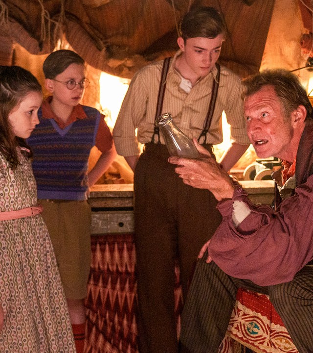 Fünf Freunde - Das Auge der Morgenröte": Der "Große Supremo" (Jason Flemyng) sitzt in einem kleinen Raum in einem Sessel. Er hält eine leere Milchflasche in der linken Hand und redet. George (Diaana Babnicova), Anne (Flora Jacoby Richardson), Dick (Kit Rakusen) und Julian (Elliott Rose) stehen um ihn herum und hören zu.