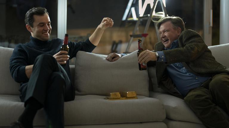 Auf einem Sofa vor einem Fenster, durch das eine nächtliche Stadtkulisse zu erahnen ist, sitzen Bernd Hausner (Justus von Dohnányi) und Sven Lebert (Nils Strunk) und prosten sich mit Bierflaschen zu, während zwischen ihnen mehrere große Goldbarren liegen.