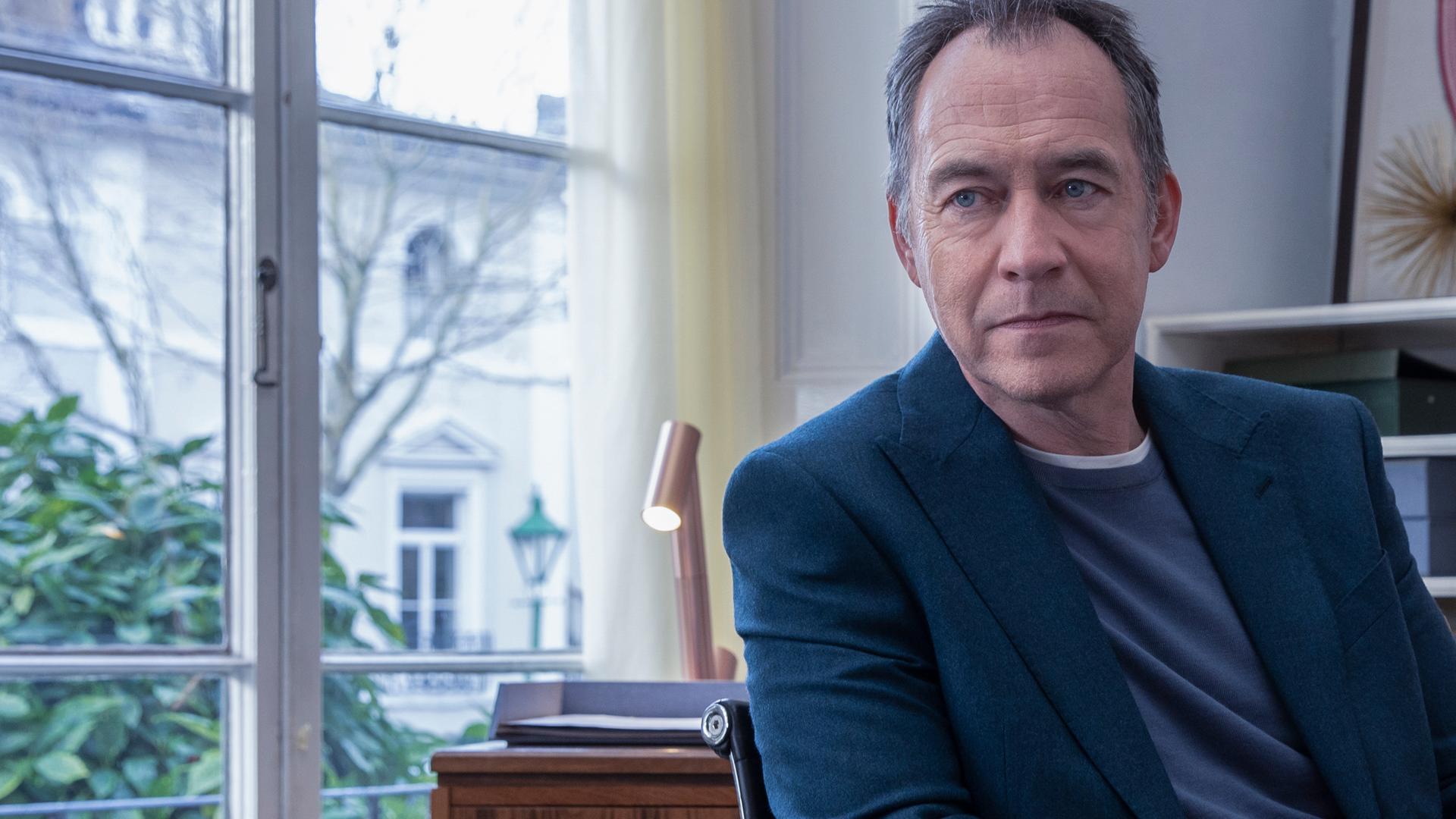 Iain Frankland (Dominic Mafham) sitzt in seinem Büro vor einem Fenster. Er trägt ein Jackett und blickt leicht zur Seite.